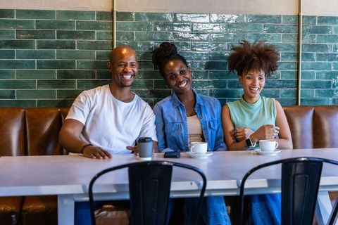 Three friends smiling and sipping coffee on leather bench with green tile backdrop