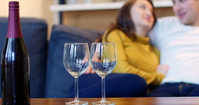 Couple sitting on couch in living room, focusing on wine glasses and bottle in foreground, suitable for themes related to relaxation, romance, companionship, and leisure time at home.