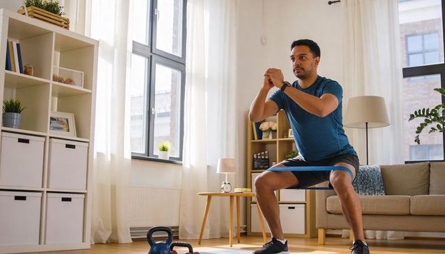 Man squatting with resistance band in bright living room doing home workout with kettlebell