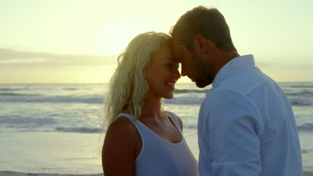 Romantic couple embracing and smiling with heads touching near the ocean during a picturesque sunset. Ideal for use in promotional materials about relationships, travel, summer holidays, and intimate moments. Can be utilized in advertisements, romantic stories, blog posts, or social media campaigns emphasizing love and connection.