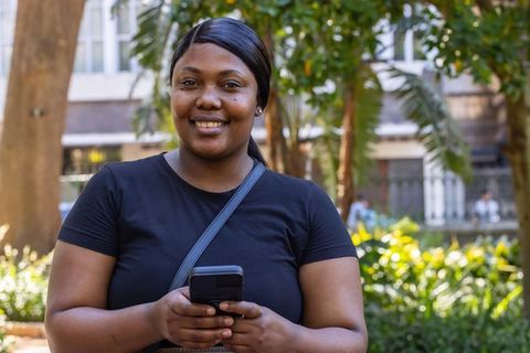 Smiling african woman holding smartphone and checking messages in sunny urban courtyard
