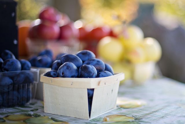 Blue ripe plums in a wooden basket are displayed on a table at a market stall, with a variety of other colorful fruits in the background. Ideal for use in materials related to healthy eating, fresh produce, farmers' markets, organic food, and seasonal harvest themes.