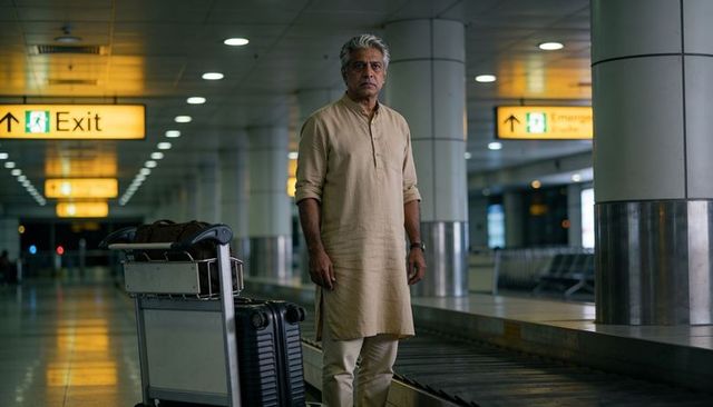 Senior indian man standing at baggage claim in beige kurta waiting with luggage trolley