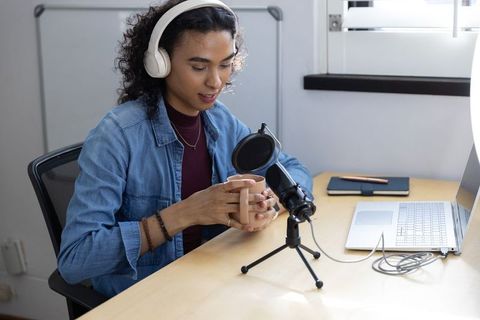 Nonbinary creator recording podcast at home desk with headphones, microphone, mug