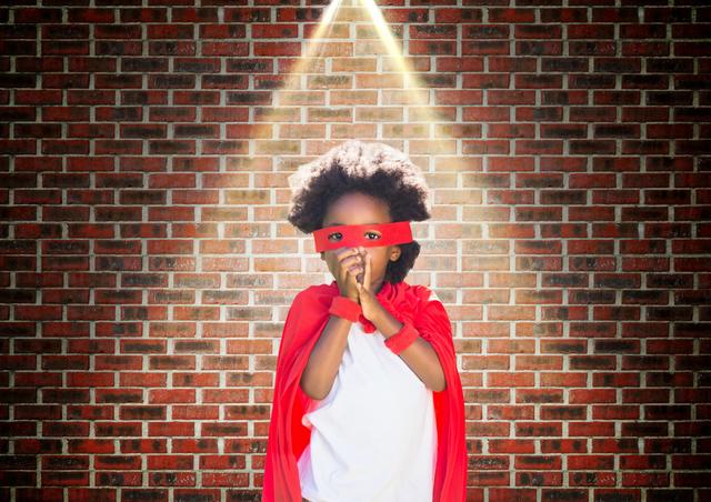 Digital composition of superhero kid in red cape and eye mask standing against brick wall