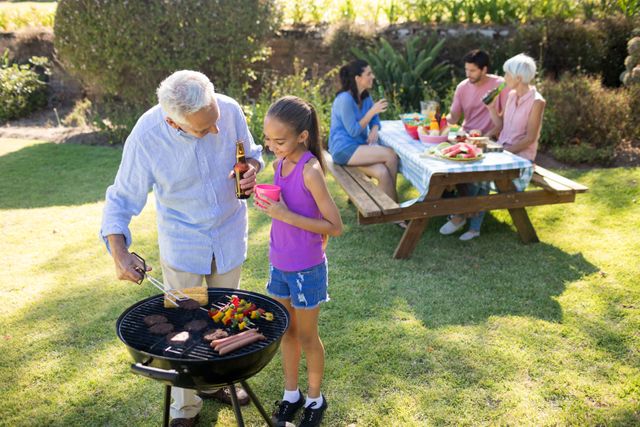 Grandfather and granddaughter grilling food on barbecue while other family members enjoy meal at picnic table in background. Perfect for illustrating family gatherings, outdoor activities, summer fun, and multigenerational bonding. Ideal for advertisements, blogs, and articles about family life, outdoor cooking, and leisure activities.