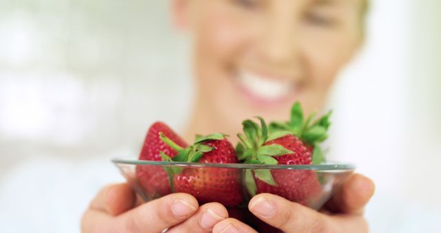 Woman holding transparent bowl filled with fresh strawberries, smiling joyfully. Ideal for topics on healthy eating, fresh produce, fruit, and wellness. Could be useful for advertisements, healthy food blogs, and lifestyle articles.