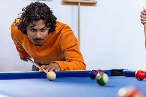 South asian man leaning and aiming cue ball on blue pool table in casual billiards game