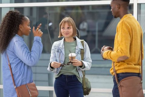 Three friends chatting over coffee outside modern glass building, casual urban fashion