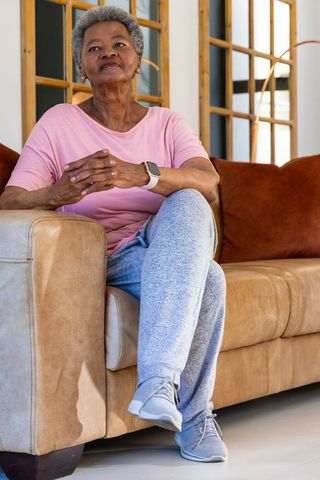 Senior woman sitting on tan leather sofa wearing smartwatch and sneakers, serene lifestyle