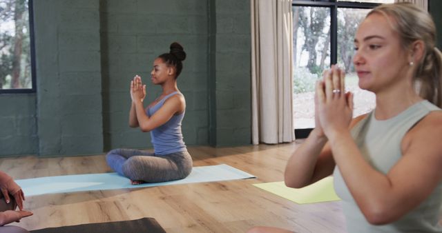 Women sitting on yoga mats, practicing yoga indoors in a meditation pose. Scene portrays relaxation, mindfulness, and physical fitness. Ideal for use in promoting wellness centers, fitness programs, yoga classes, health magazine articles, or lifestyle blogs focused on fitness and mental well-being.