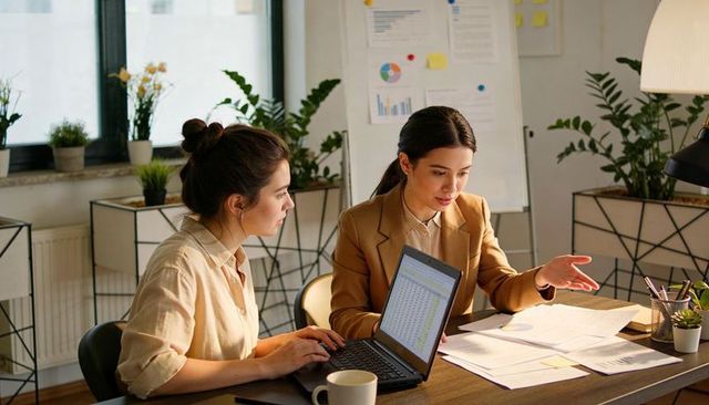 Two professional women collaborating over laptop and documents in modern office workspace