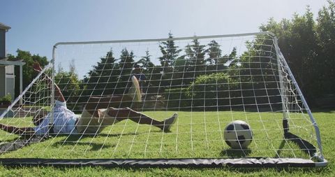 African american mentor coaching teenage boy in backyard soccer, goalkeeper diving save