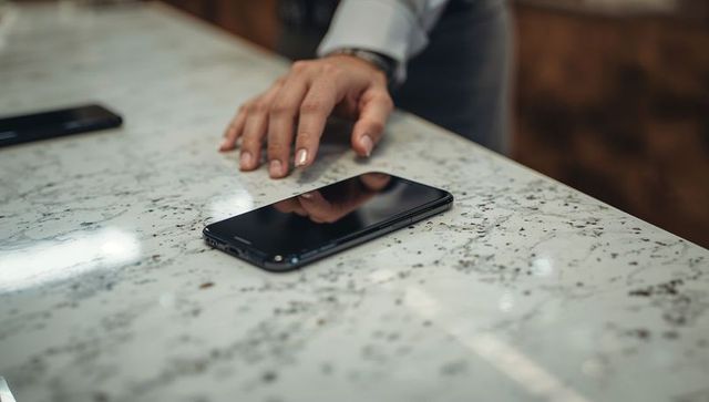 Manicured hand reaching glossy smartphone on polished stone countertop with reflective bokeh