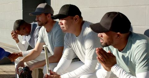 Sunlit multiethnic baseball teammates sitting on dugout bench holding bat helmet glove