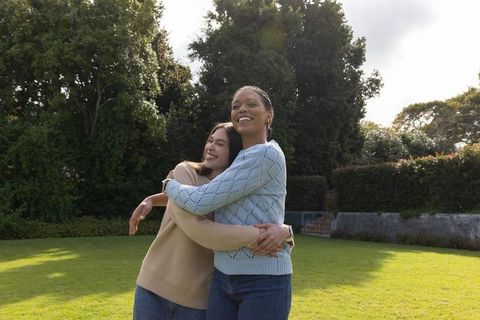 Diverse female couple embracing and smiling on sunlit backyard lawn wearing knit sweaters