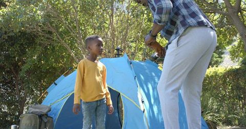 Man drilling to secure backyard tent while boy watching during sunny garden camping
