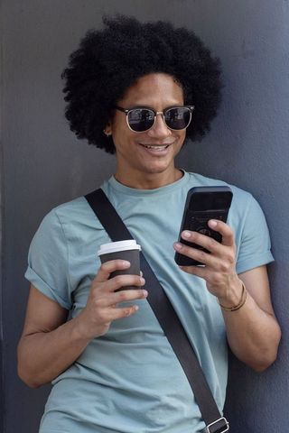Man leaning on gray wall checking smartphone, holding coffee cup, wearing sunglasses