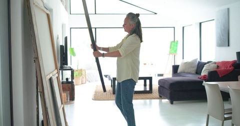 Mature woman positioning large framed canvas in bright home art studio living room