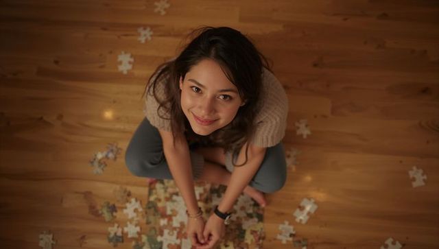 Smiling young woman assembling jigsaw puzzle on hardwood floor, cozy warm at-home scene