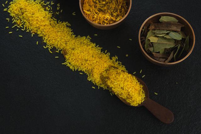 Close-up view of yellow rice and various spices on a black background. The image features a wooden scoop filled with yellow rice, and bowls containing bay leaves and other spices. Ideal for use in culinary blogs, recipe websites, cooking magazines, and food-related advertisements to highlight ingredients and cooking themes.