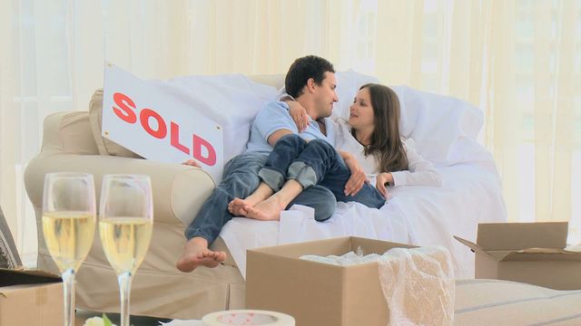 Couple embracing on a comfortable sofa amidst unboxed belongings, expressing joy in their new home with a sold sign prominently placed. Perfect for concepts of new beginnings, property ownership, or real estate marketing.