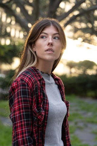 Young woman standing in park at golden hour wearing red plaid shirt and scalloped gray top