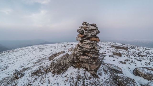 Stone cairn marking windswept alpine summit dusted with snow and lichen, rugged panorama