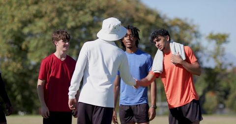Coach greeting diverse young male athletes on sunny outdoor field, teamwork and fitness