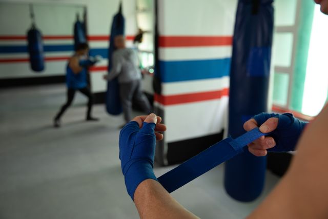 Boxer wrapping hands with blue wraps in a gym while another boxer is sparring in the background. Ideal for use in articles or advertisements about fitness, boxing training, sports preparation, and athletic routines.