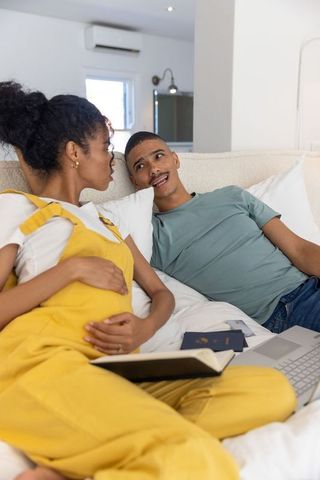 Couple relaxing on bed, reading and planning trip with laptop and passport