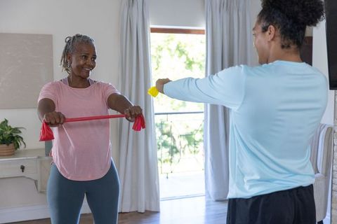 Senior woman and coach exercising with resistance band and dumbbell in sunlit living room