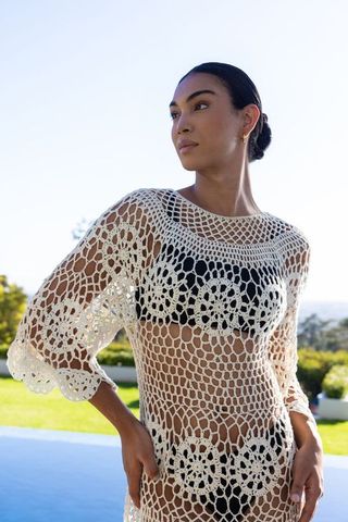 African woman wearing white crochet coverup over black swimsuit poolside overlooking ocean