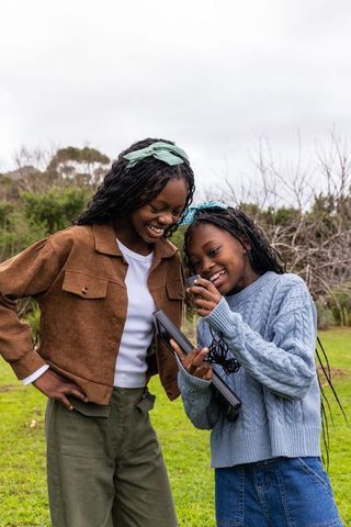 African american teen girls laughing and exploring tablet outdoors in meadow