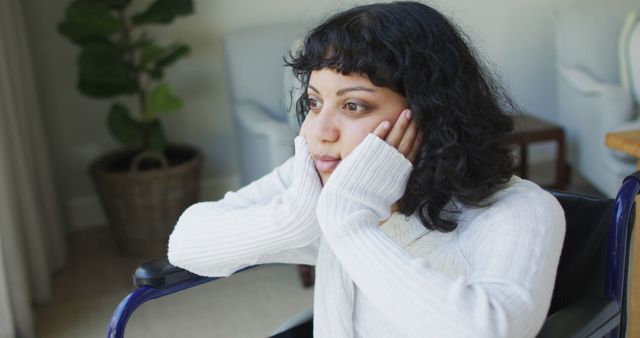 A young woman with curly hair is sitting in a wheelchair indoors, looking contemplative. Her hands are resting on her face, enhancing her pensive expression. She appears to be lost in thought while gazing into the distance. Suitable for themes of disability, contemplation, mental health, personal reflection, and lifestyle imagery.