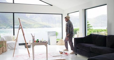 Indian artist walking toward window in sunlit lakeside studio with easel and paints