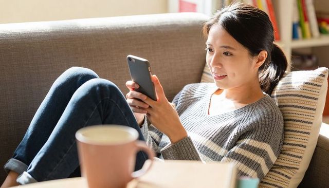 Asian woman relaxing on sofa, smiling while using smartphone with coffee and books