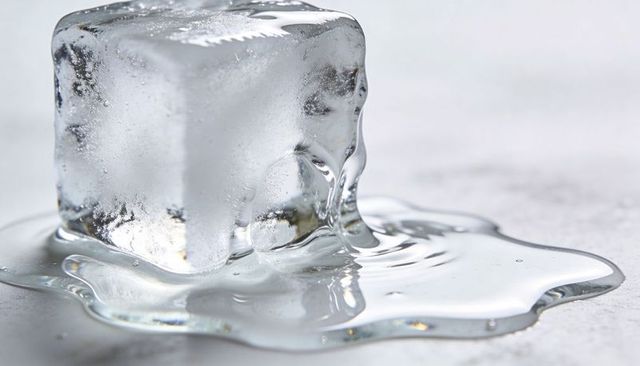Melting crystal-clear ice cube forming glossy water puddle with bubbles and reflections