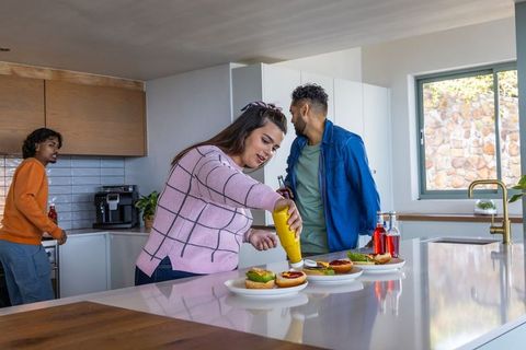 Friends making burgers on modern kitchen island, woman squeezing mustard during casual gathering