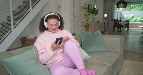 African american woman relaxing on couch in pink loungewear with headphones using smartphone