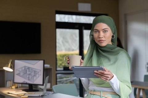 Female architect wearing green hijab holding tablet and mug in modern office with 3d plan