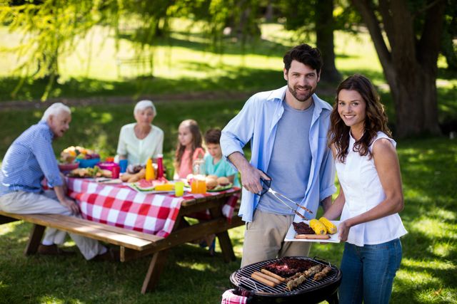 Family enjoying outdoor barbecue picnic in park. Smiling couple grilling food while family members sit at picnic table. Ideal for promoting family gatherings, outdoor activities, summer events, and leisure time.