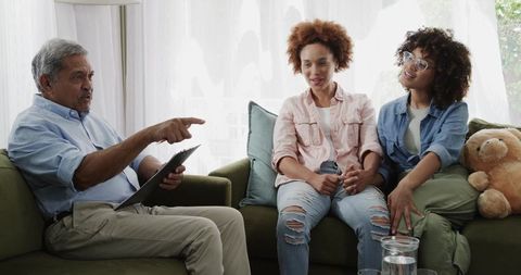 Senior man pointing tablet while adult daughters holding hands on couch with teddy bear