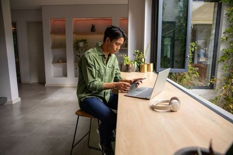Asian man working remotely at wooden counter using laptop and smartphone by window nearby