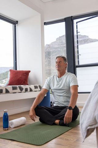 Mature man meditating on green yoga mat by sunlit window with mountain view for wellness