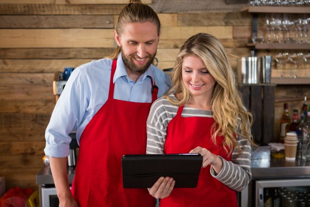 Two baristas in a coffee shop are using a digital tablet, both smiling and wearing red aprons. The background features a wooden wall and various coffee shop equipment. This image can be used for promoting coffee shops, teamwork, small businesses, and the use of technology in customer service.