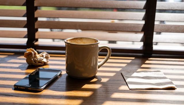 Beige ceramic mug catching warm morning sunlight through wooden blinds on cafe table