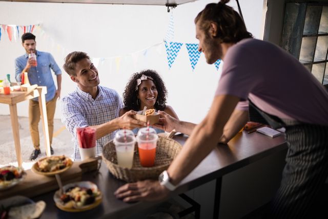 Waiter serving snacks to smiling customers at a food truck counter. The scene includes colorful drinks and food items on the counter, with a festive atmosphere created by hanging decorations. Ideal for use in marketing materials for street food festivals, outdoor events, or food truck businesses.