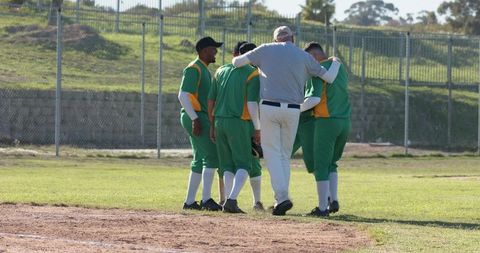 Coach supporting struggling player while teammates huddling on community baseball field