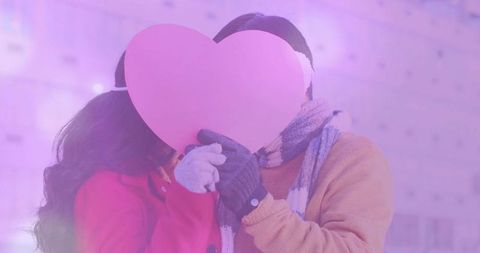 Couple holding pink heart while wearing winter coats and scarves, romantic city portrait